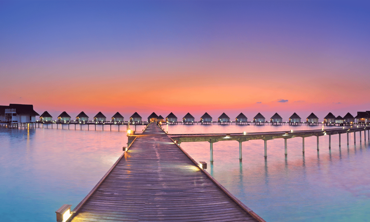 Holzsteg führt zu Wasserbungalows im Meer unter einem farbenprächtigen Sonnenuntergang. Lichter spiegeln sich im ruhigen Wasser.