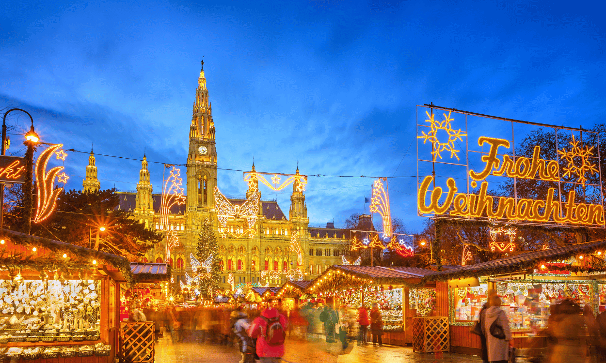 Weihnachtsmarkt vor dem hell erleuchteten Wiener Rathaus bei Dämmerung, mit geschmückten Buden und Leuchtschrift Frohe Weihnachten